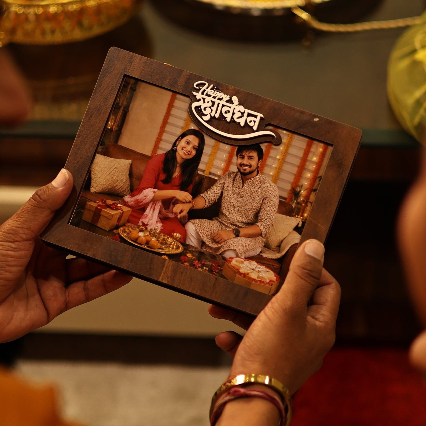 A person holds a small wooden shadow box frame containing a family photo and a decorative script label. The background is warmly lit and shows other decorative items.
